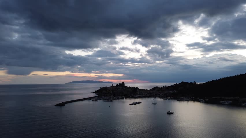 Aerial view of coastal town with historic castle and harbor at sunset over the Mediterranean Sea with dramatic clouds Italy.