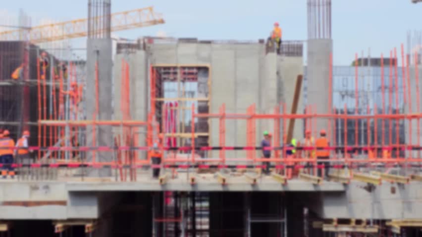 Construction site with steel beams and concrete framework. Workers wear orange safety vests. They work closely together on scaffolding. Cranes lift materials in the background
