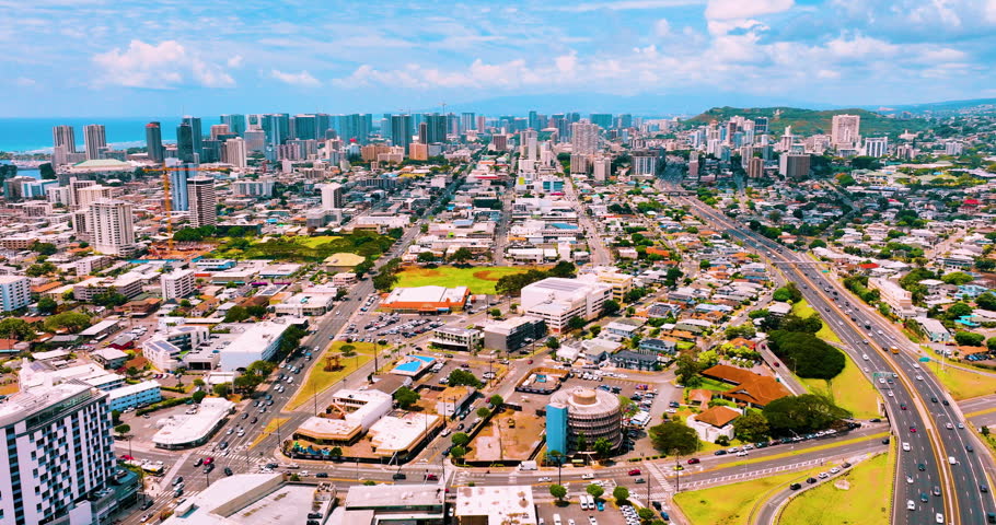 Aerial view of Honolulu urban grid and downtown skyline, Hawaii. Street network, mixed residential and commercial district, ocean and green hills in distance under partly cloudy sky.