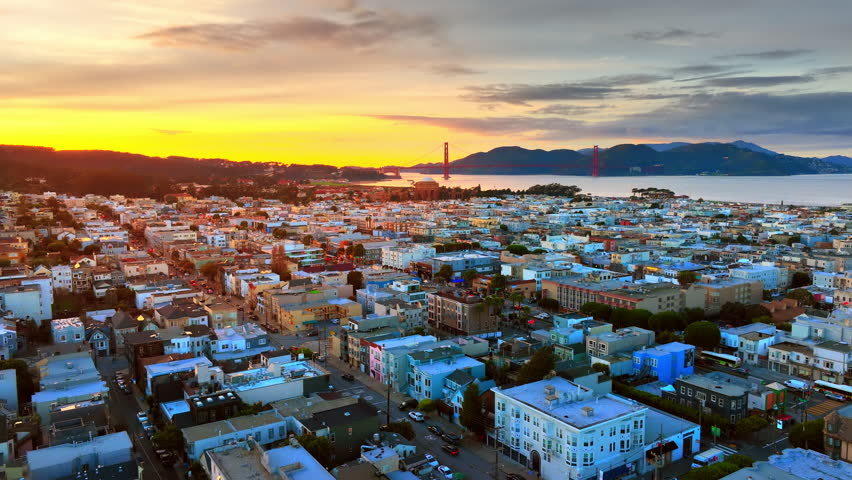 Sunset glow over San Francisco Richmond neighborhood toward Golden Gate Bridge and Marin headlands. Dense pastel residential grid, iconic red bridge tower, bay and mountain silhouette.