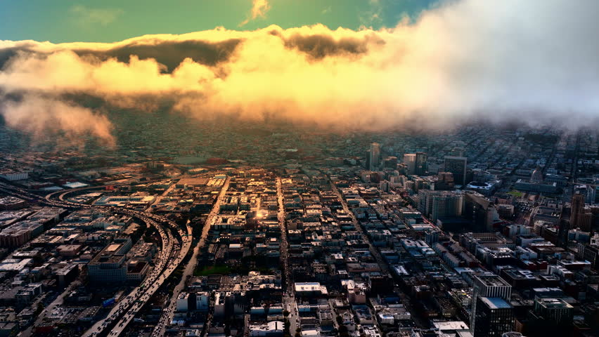 Low cloud layer rolling over urban grid at dusk, San Francisco. Golden lit fog bank descending on flat city blocks, freeway curve at left, teal and amber sky above.