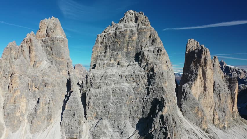 Spectacular close-up drone shot over the Italian Dolomites. Features towering, rugged grey limestone mountain peaks and jagged rock formations standing majestic under a clear blue sky.