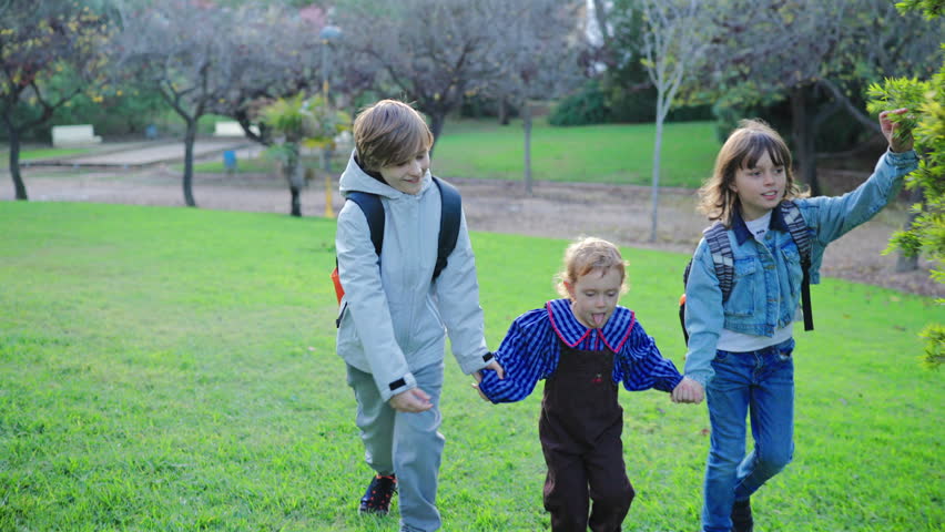 Three happy children holding hands while running together across lush green grass in a park. Two older kids with backpacks guide a younger toddler in overalls through the sunny outdoor space.