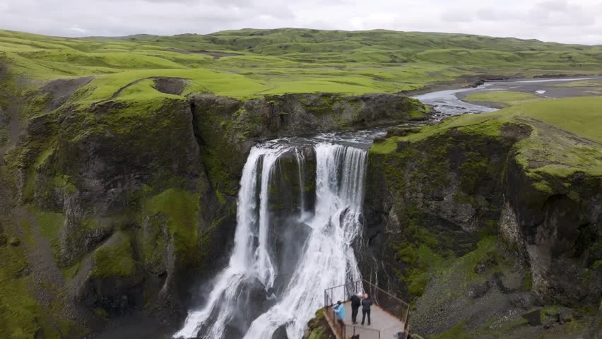 Aerial view of a powerful waterfall cascading over mossy cliffs with tourists standing on a wooden observation deck in Iceland.