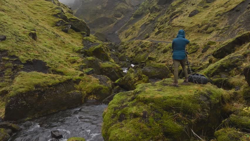 View of a photographer standing on a mossy rock formation with a tripod, capturing footage of a stream flowing through a rugged, green valley in Iceland.