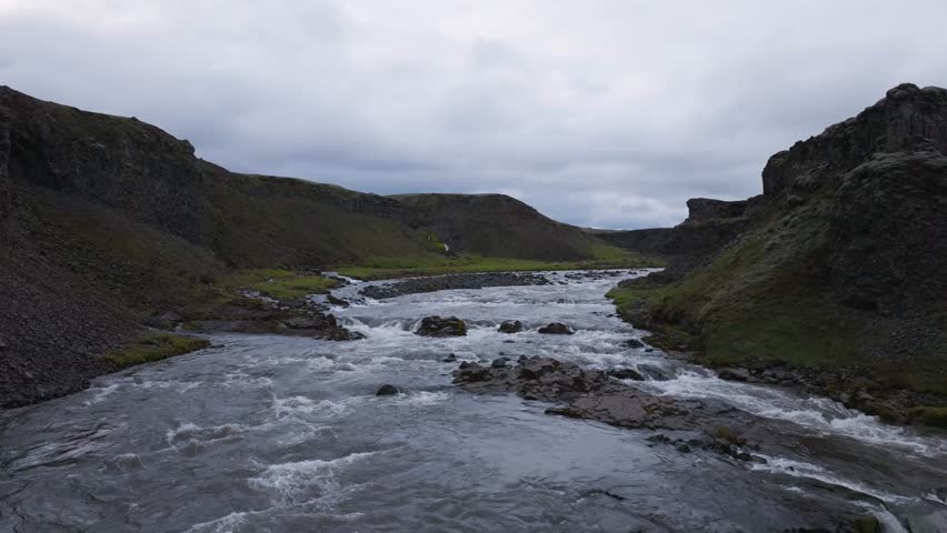 Aerial view of a river flowing through a rocky landscape with green hills and dark volcanic rocks under a cloudy sky in Iceland.