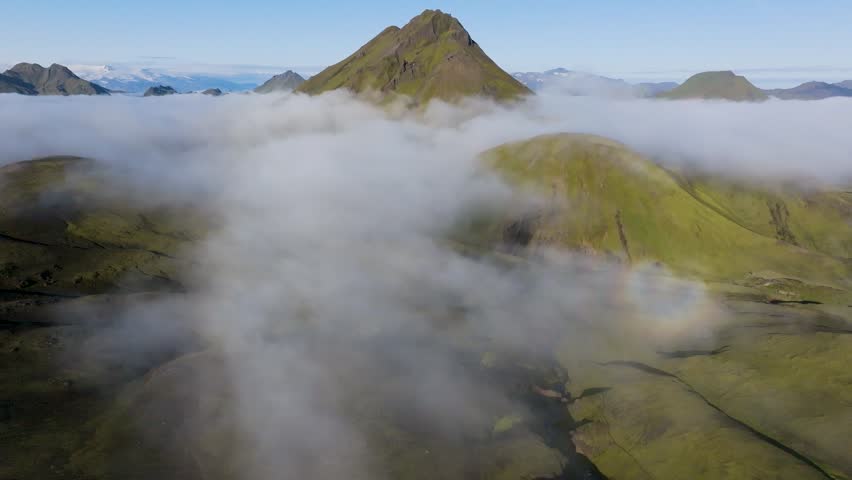 Aerial view of green mountains rising above a thick layer of low-hanging clouds under a clear blue sky in the highlands of Iceland.