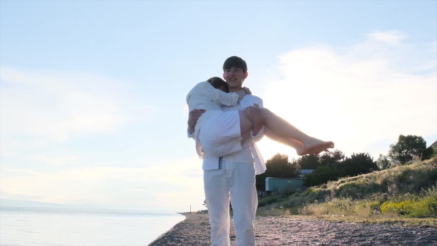 Teenage boy carrying a girl in his arms on a seashore at sunset. Young couple, first love and sibling relationship concept as happy teenagers enjoy time together by the sea in white clothes.
