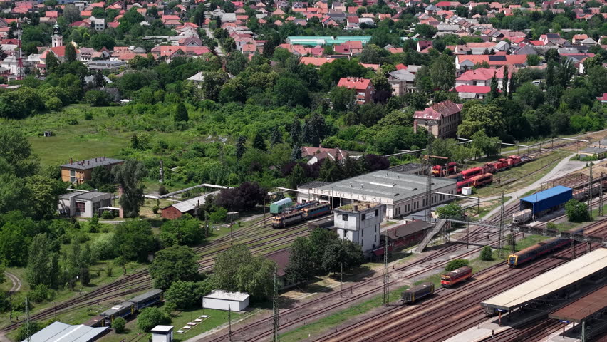 An aerial perspective captures a functional train station depot nestled within a residential town in Hungary during a summer day.
