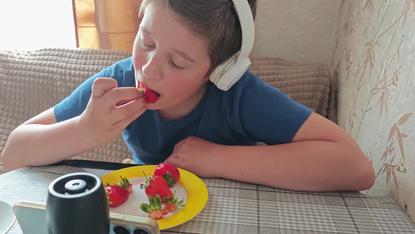 Young Caucasian boy wearing headphones enjoys fresh strawberries while seated at a table, with a plate of strawberries and a black speaker visible in the cozy indoor setting