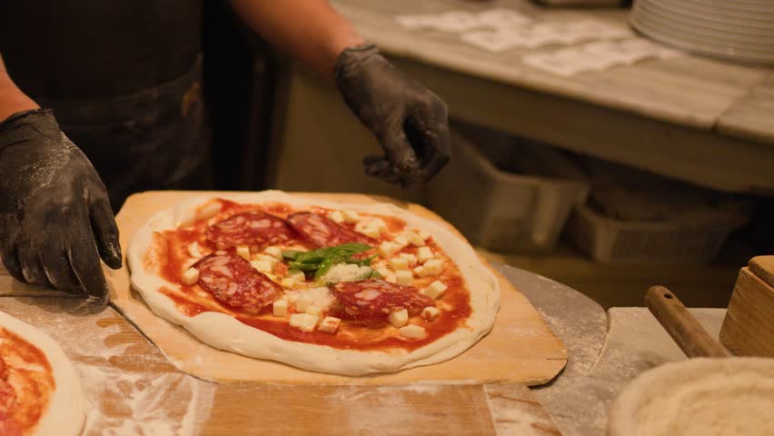 Close-up of a chef in black gloves lifting a raw pizza with a wooden peel.