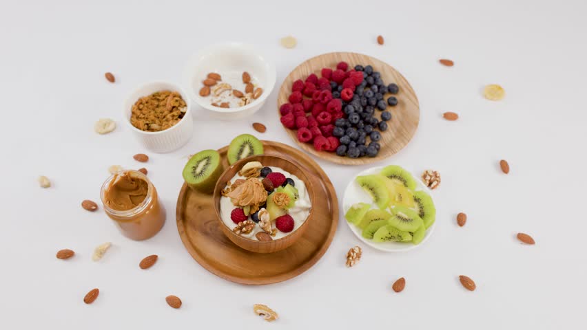 Overhead shot of hands presenting a yogurt bowl with berries, kiwi, and nuts on white.