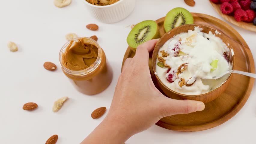 Overhead shot of hands scooping yogurt from a wooden bowl filled with berries and nuts.