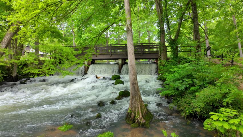 View of the cascading waters of Maramec Spring Park flowing under a wooden bridge surrounded by lush green trees and mossy rocks in Saint James, Missouri, United States.