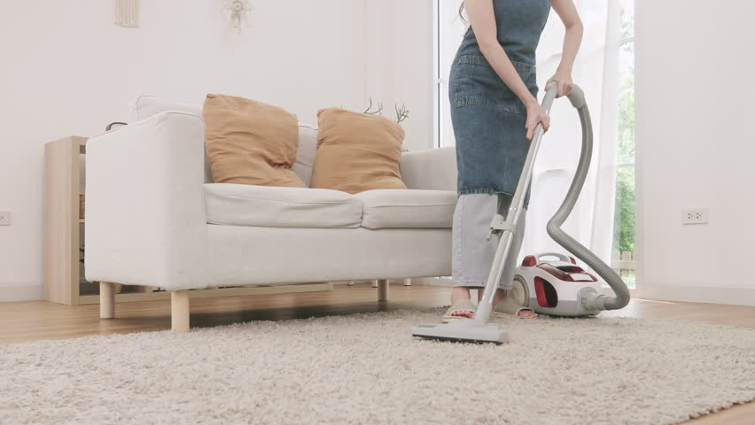 A woman using a canister vacuum cleaner to clean a fluffy carpet in a bright and cozy living room.