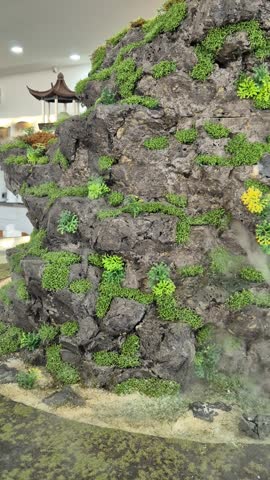 Mossy rock wall with lush green plants and mist rising from base. Yellow flowers peek through dense foliage for contrast. Stone waterfall cascades softly over rugged terrain