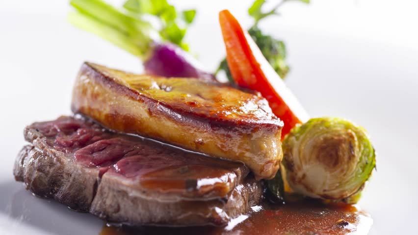 Luxury French steak topped with seared foie gras and roasted vegetables on dark plate revealed by slow panning camera
