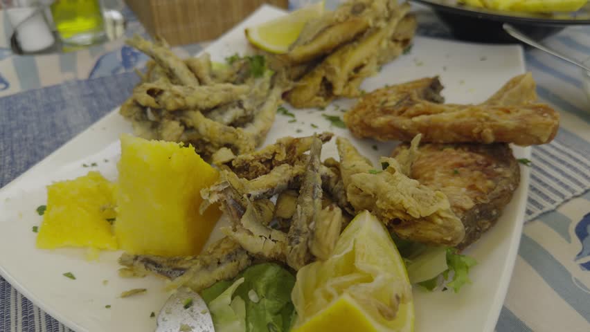 Delicious mix of crispy fried seafood served with a slice of lemon and traditional polenta on a white plate. Close up of golden battered fish assortments cooked for a fresh Mediterranean lunch. Traditional tavern meal offers a variety of crunchy fried small fish options. This tasty seafood platter is served on a restaurant table for a authentic culinary dinner. Food lovers enjoy a hot gourmet seafood recipe