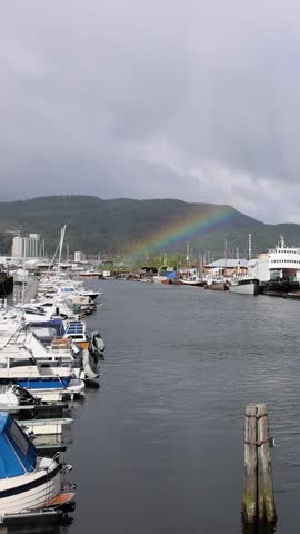 View of numerous boats moored in the harbor with a rainbow appearing over the forested hills under a cloudy sky in Trondheim, Trondelag, Norway.