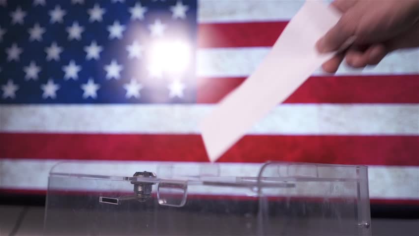 Hand putting a ballot paper into a transparent ballot box on the background of the USA flag. Concept of voting, democracy and upcoming US Senate elections.