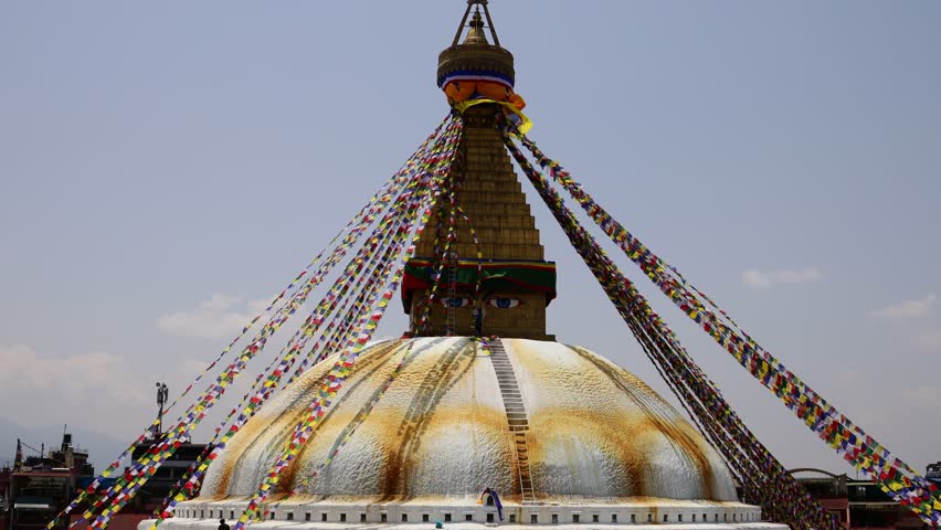 Video of Boudhanath Stupa in Kathmandu, Nepal, one of the most important Buddhist monuments in the world. The massive white dome and iconic gilded spire, adorned with the Buddha eyes, stand at the center of a vibrant religious and cultural complex. Colorful prayer flags stretch across the scene, fluttering in the wind and adding movement and atmosphere to the footage. Pilgrims and visitors often circle the stupa in a traditional act of devotion, contributing to the spiritual character of the location. Recognized as a UNESCO World Heritage Site, Boudhanath is a major center of Tibetan Buddhism and an important landmark in Nepal. The video captures the architecture, religious traditions, and cultural heritage of Kathmandu, making it suitable for travel, spirituality, religion, culture, and documentary projects