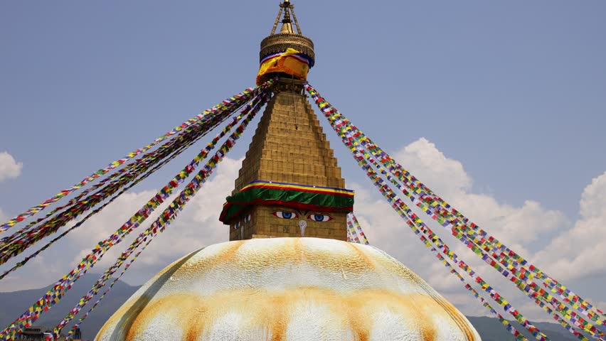 Video of Boudhanath Stupa in Kathmandu, Nepal, one of the most important Buddhist monuments in the world. The massive white dome and iconic gilded spire, adorned with the Buddha eyes, stand at the center of a vibrant religious and cultural complex. Colorful prayer flags stretch across the scene, fluttering in the wind and adding movement and atmosphere to the footage. Pilgrims and visitors often circle the stupa in a traditional act of devotion, contributing to the spiritual character of the location. Recognized as a UNESCO World Heritage Site, Boudhanath is a major center of Tibetan Buddhism and an important landmark in Nepal. The video captures the architecture, religious traditions, and cultural heritage of Kathmandu, making it suitable for travel, spirituality, religion, culture, and documentary projects