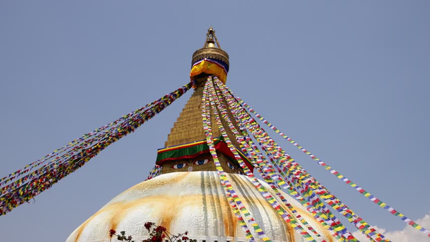 Video of Boudhanath Stupa in Kathmandu, Nepal, one of the most important Buddhist monuments in the world. The massive white dome and iconic gilded spire, adorned with the Buddha eyes, stand at the center of a vibrant religious and cultural complex. Colorful prayer flags stretch across the scene, fluttering in the wind and adding movement and atmosphere to the footage. Pilgrims and visitors often circle the stupa in a traditional act of devotion, contributing to the spiritual character of the location. Recognized as a UNESCO World Heritage Site, Boudhanath is a major center of Tibetan Buddhism and an important landmark in Nepal. The video captures the architecture, religious traditions, and cultural heritage of Kathmandu, making it suitable for travel, spirituality, religion, culture, and documentary projects