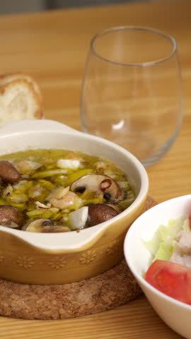 Seafood and mushroom ajillo on a dinner table with pouring white wine into a glass, accompanied by baguette and salad, vertical format.