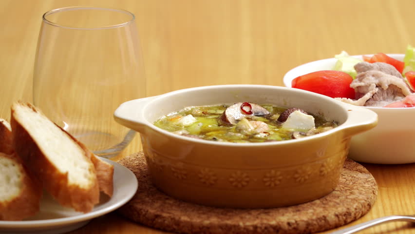 Seafood and mushroom ajillo on a dinner table with pouring white wine into a glass, accompanied by sliced baguette and salad.
