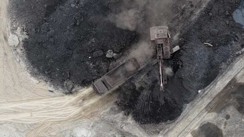 An excavator loads a dump truck with coal at a dusty industrial mining site in Hungary, illustrating heavy industry and resource extraction.