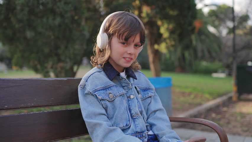 Kid on the phone listening to music while sitting on wooden park bench wearing white headphones and denim jacket. Golden hour sunlight creates warm atmosphere in urban park setting.