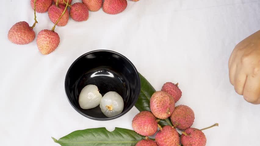 A woman hand Holding a Freshly Peeled Lychee Fruit, Revealing Its Juicy White Flesh and red Outer Shell on keep black bowl. inside with another whole lychee or litchi fruits.