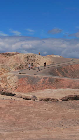 Vertical Screen: Tourists explore colorful geological formations at Zabriskie Point in Death Valley National Park California with winding paved pathway through multicolored badlands under blue sky.