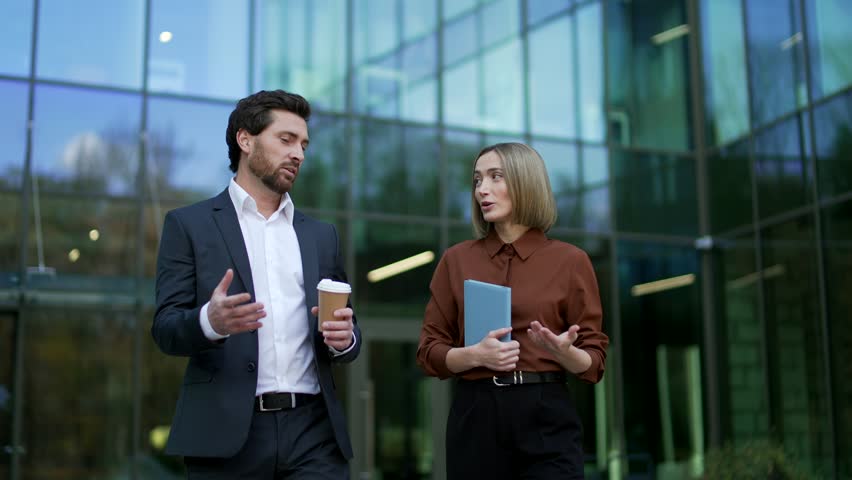 Front view close-up of smiling business colleagues with coffee cups standing near a modern glass office building. Professionals pause after a short walk and continue discussing ideas in urban outdoor