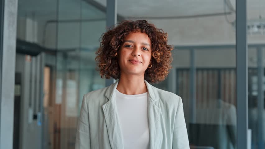 Portrait of smiling young multiracial latin or indian businesswoman with curly hair standing in office, wearing casual blazer in professional workspace. Arabian young woman girl smiling at camera