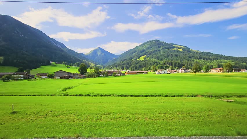 Wide Austrian countryside view from a moving train, featuring lush green fields, rural homes, forested ridges, and blue sky over a peaceful alpine valley.