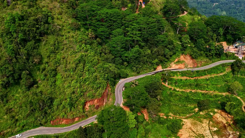 Traffic follows a winding mountain road as it curves around steep, tree-covered slopes and terraced hillsides in daylight.
