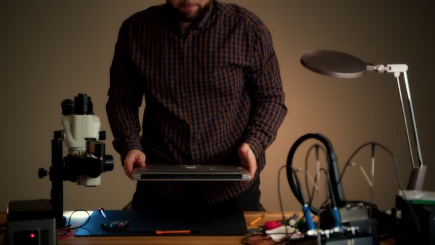 Technician repairing a laptop at a workbench with focused hand movements in a warm-lit electronics workspace. Tools and lab equipment surround him during troubleshooting