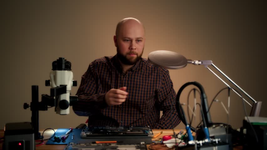 A technician in a checkered shirt inspects an open laptop at a cluttered electronics workbench. Warm workshop lighting and precision tools create a focused repair scene