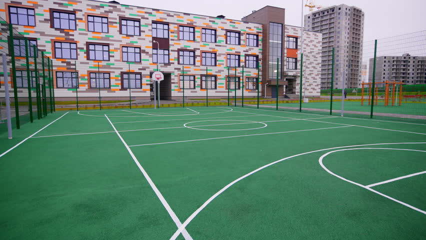 Outdoor basketball court features green surface and white markings. Modern school building with colorful facade stands under overcast sky