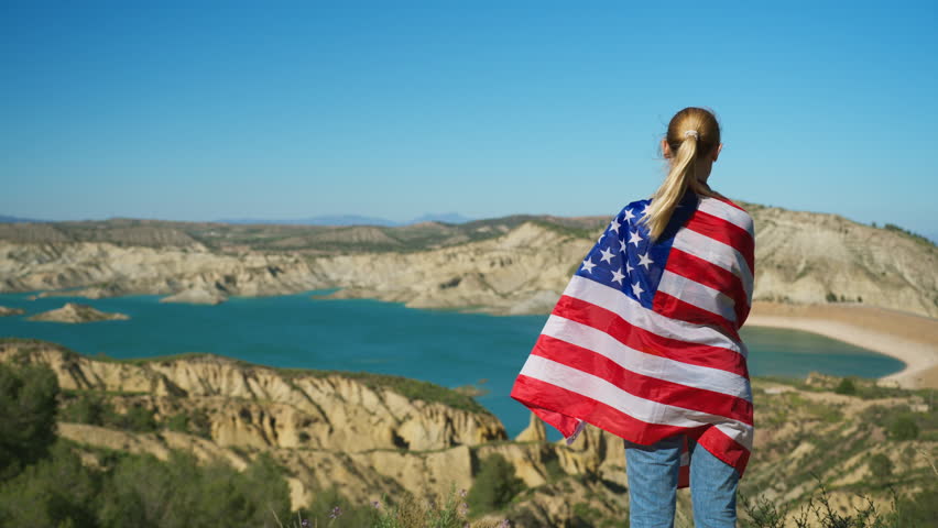 Young woman wrapped in American flag admiring breathtaking canyon and lake view. Concept of patriotism, freedom, national pride, travel, outdoor adventure and the American spirit