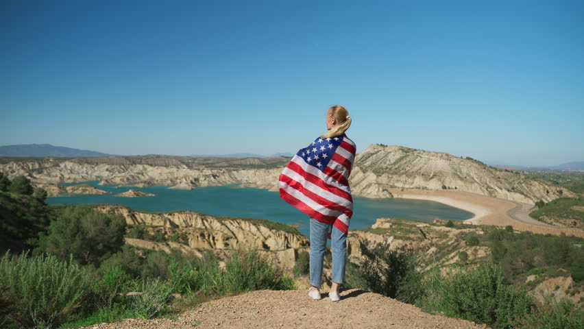 Woman wrapped in American flag enjoy peaceful moment above canyon lake. Inspiring concept of freedom, liberty, independence, adventure, travel, and personal achievement