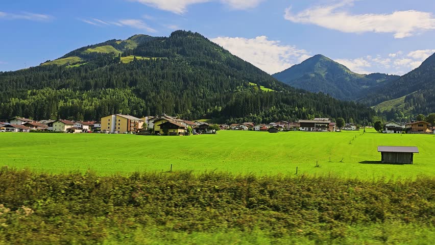 Austria railway journey through a bright alpine valley, passing grassy meadows, farmhouses, wooded slopes, and mountain peaks under soft clouds and clear summer sky.