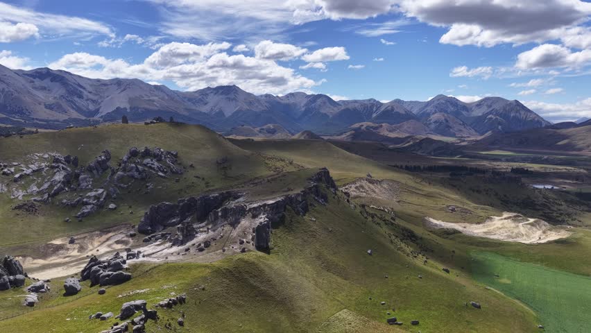 Aerial drone footage over the Southern Alps of New Zealand South Island showing dramatic mountain peaks rocky cliffs green valleys and a winding river under a blue cloudy sky.