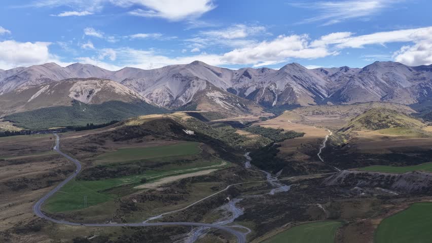 Aerial drone footage over an arid alpine valley in New Zealand with golden tussock grasslands rocky mountains a winding road and river under a dramatic blue sky.
