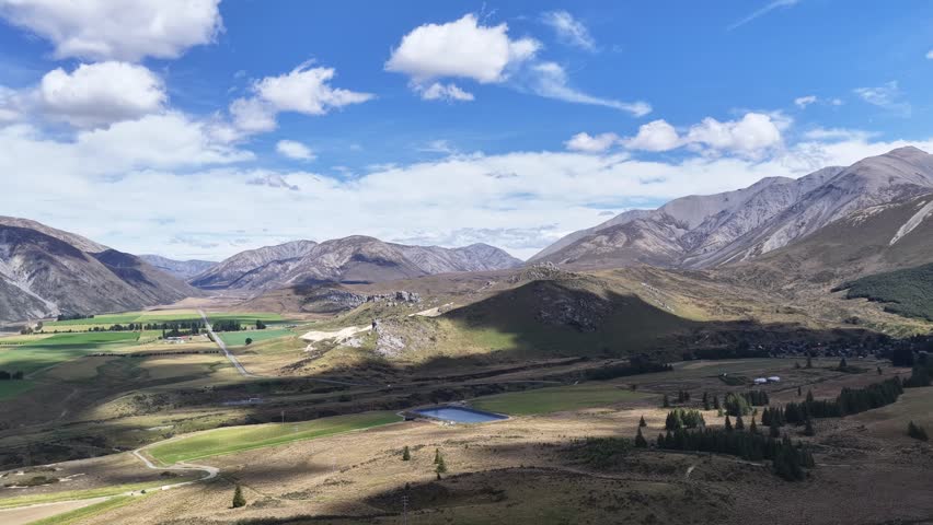 Aerial drone footage over a wide alpine valley in New Zealand with green farmland tussock grasslands rocky mountains and snow capped peaks under a blue sky.