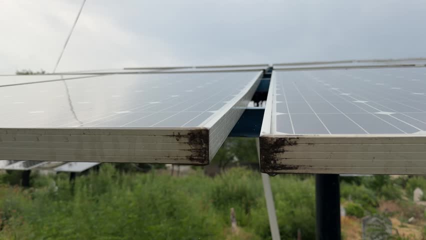 Solar panels installed in a graveyard overlook a lush landscape, rainfall cascading under heavy, dark clouds.