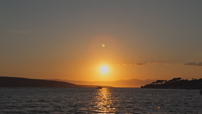 Static wide shot of a beautiful golden sunset over a calm sea bay, with small fishing boats anchored near a silhouetted island on a peaceful summer evening.