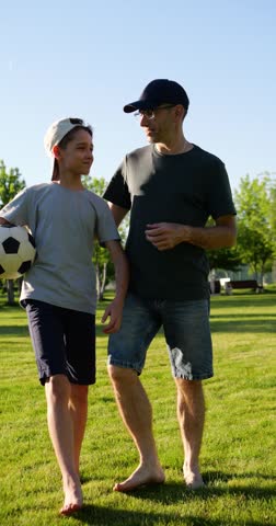 Father and son walking barefoot through park at sunset while talking and enjoying time together. Boy carries soccer ball as golden evening light highlights family connection and shared moments.
