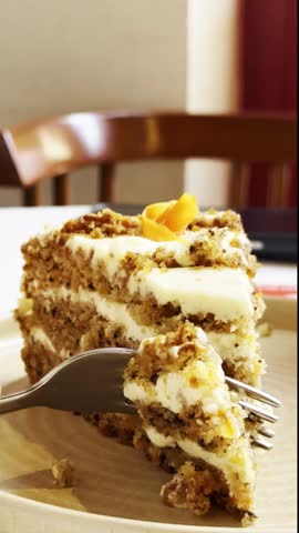 A slice of carrot cake on a plate with a fork. The cake has layers of cream cheese frosting and is topped with a small piece of carrot. The background features a wooden chair.